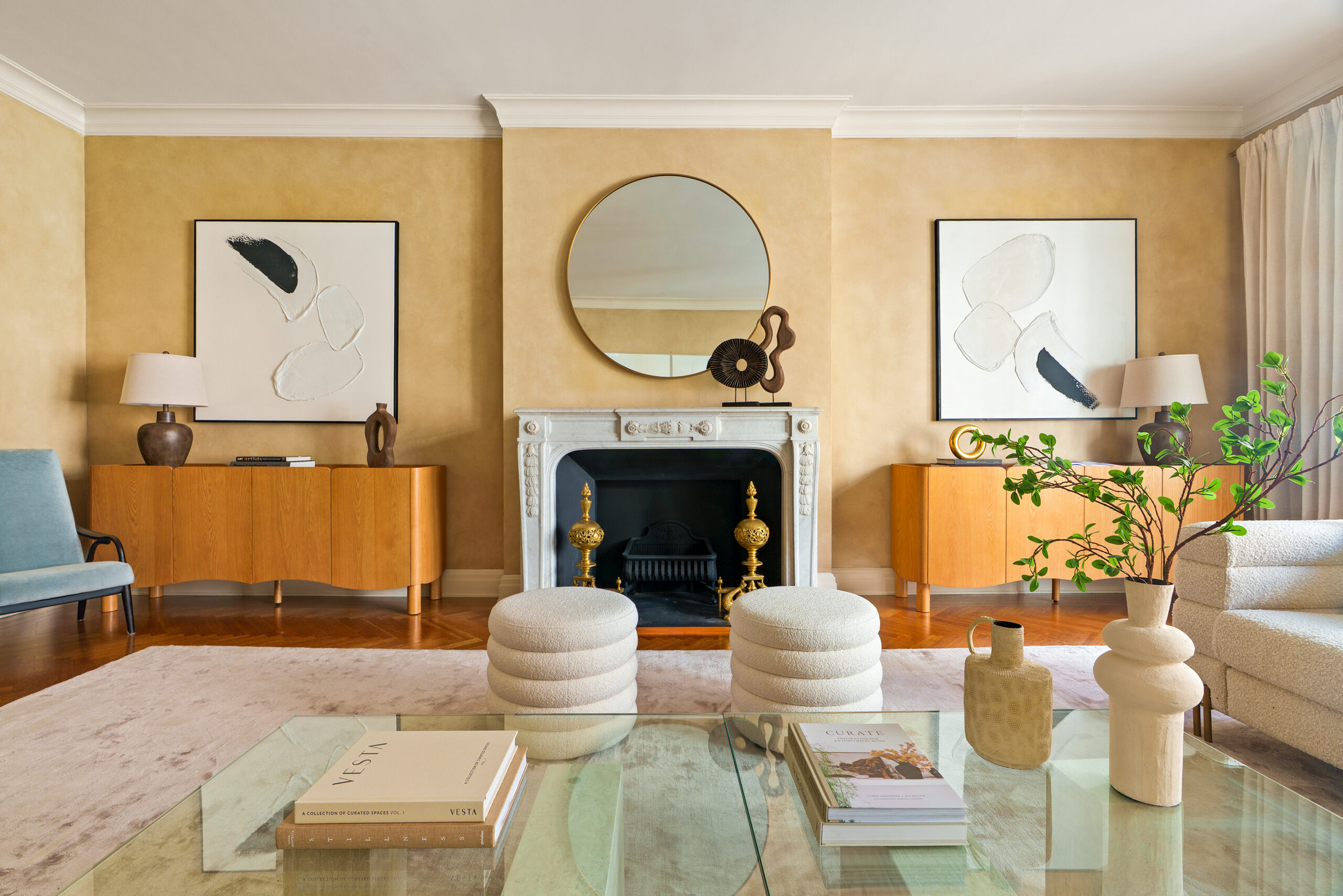 Living room with a fireplace, two white ottomans, matching wooden consoles topped with abstract art, and a glass coffee table with books.