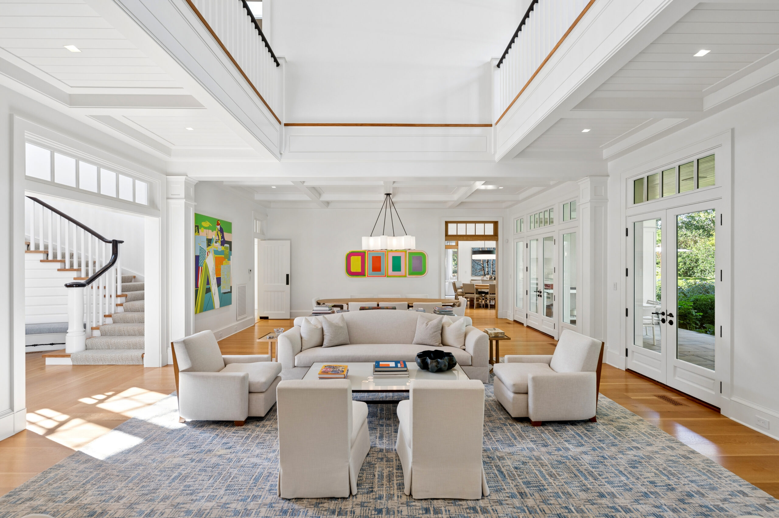 A living room with a white sofa and four armchairs arranged around a coffee table under a high ceiling with a central chandelier, featuring a colorful abstract painting on the wall.
