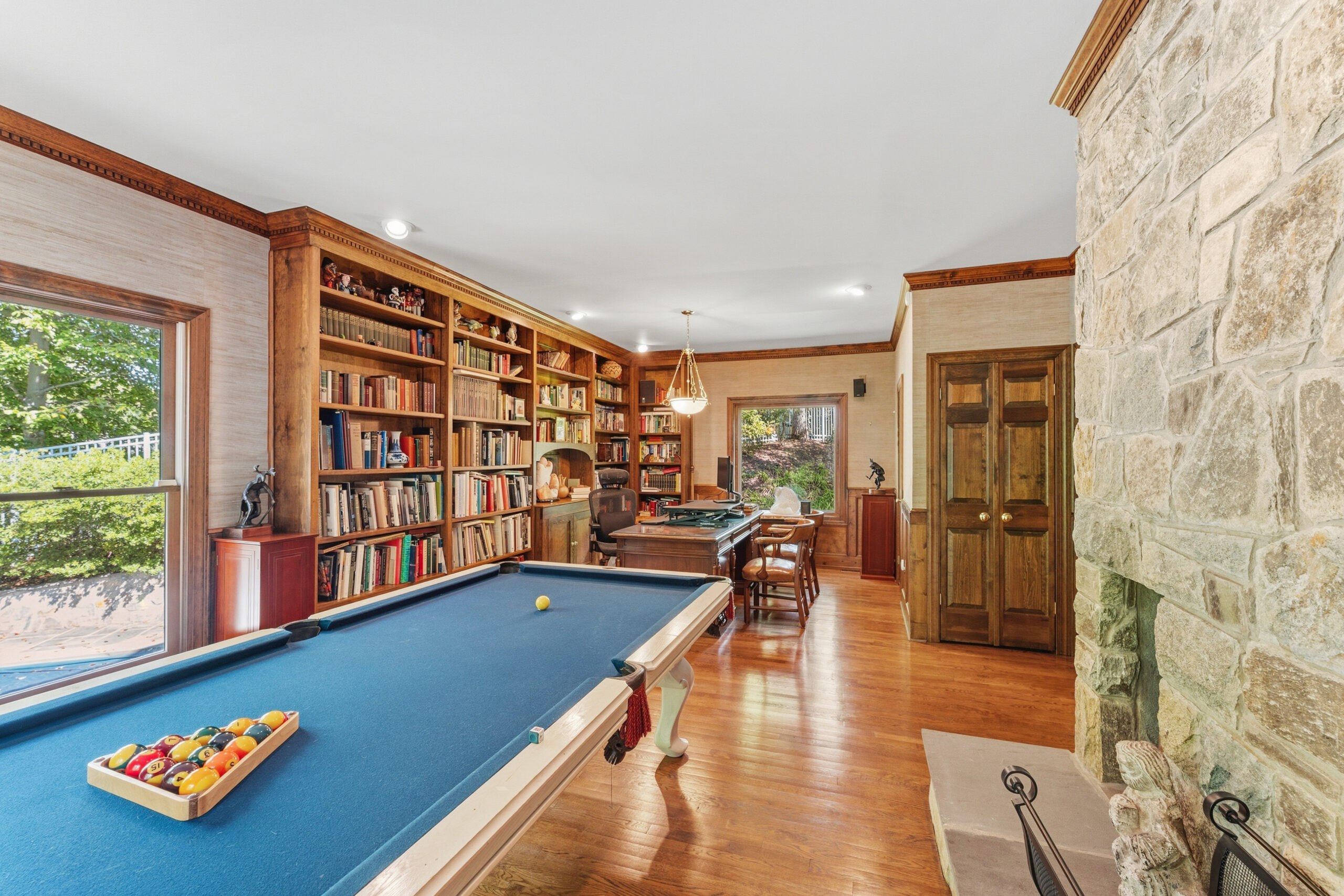 708 Potomac Knolls A home office features a blue pool table in the foreground, floor-to-ceiling wooden bookshelves, a desk with chairs, and a large stone fireplace.