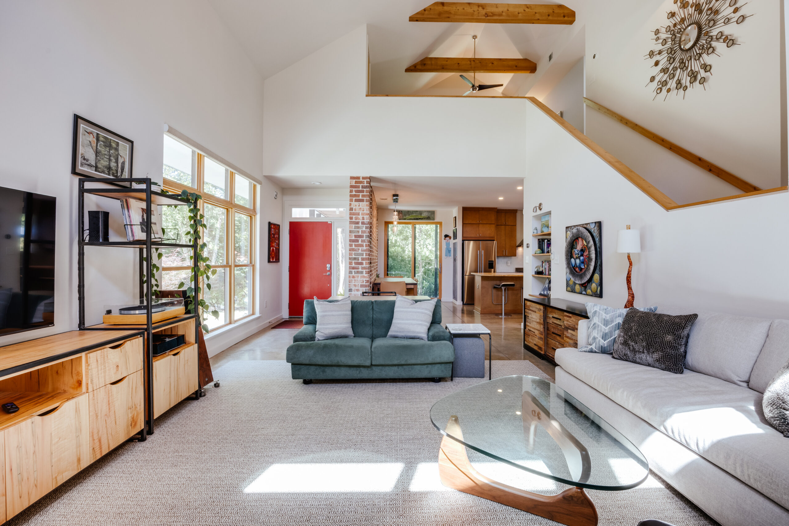 Open-concept living room from November 2026 with a green sofa facing a television console, a light gray sofa with a glass coffee table, and a lofted area above a kitchen in the background.