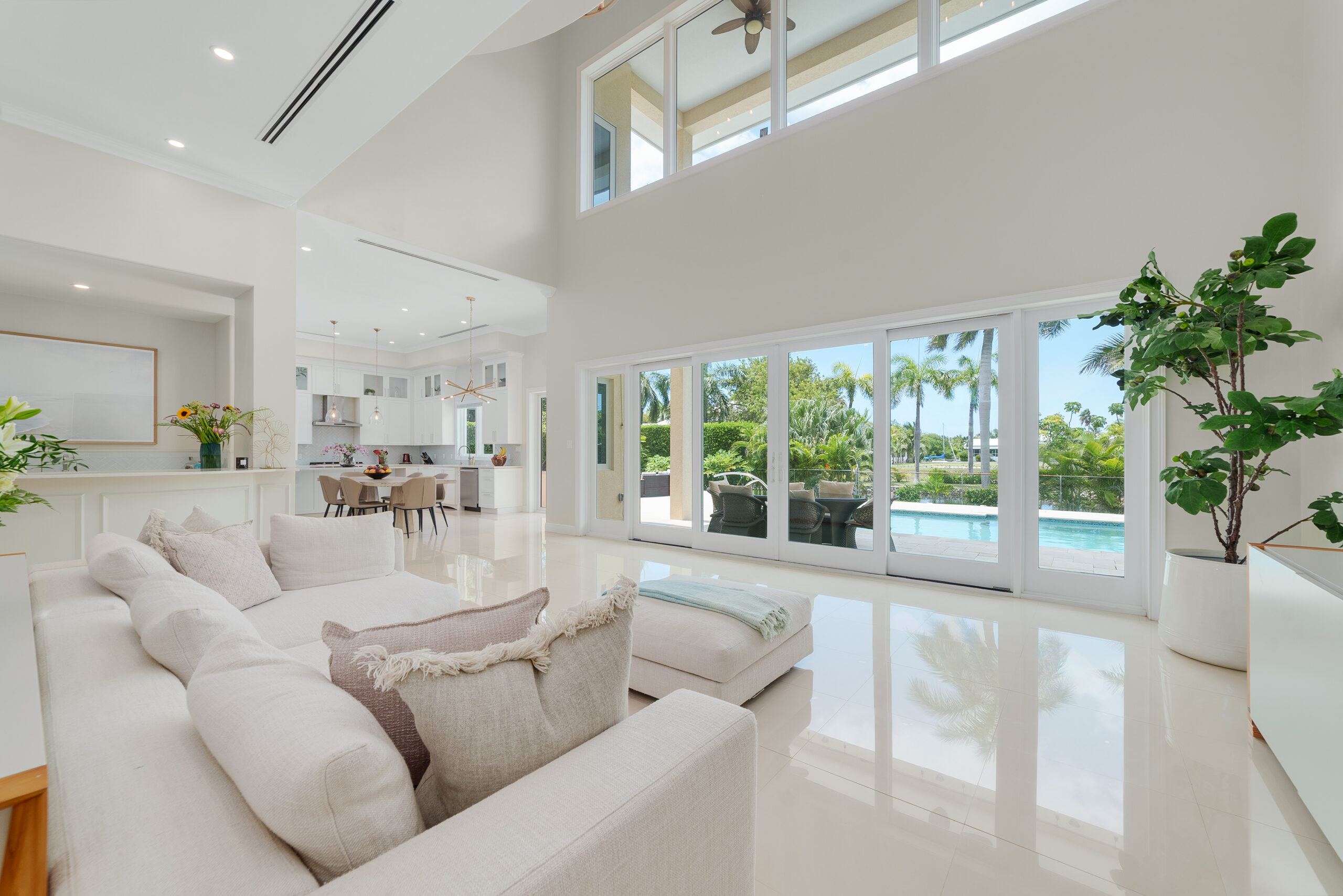 A bright, open-plan living room with white sofas looking out through floor-to-ceiling glass doors toward a swimming pool and patio, ideal for November 2026.