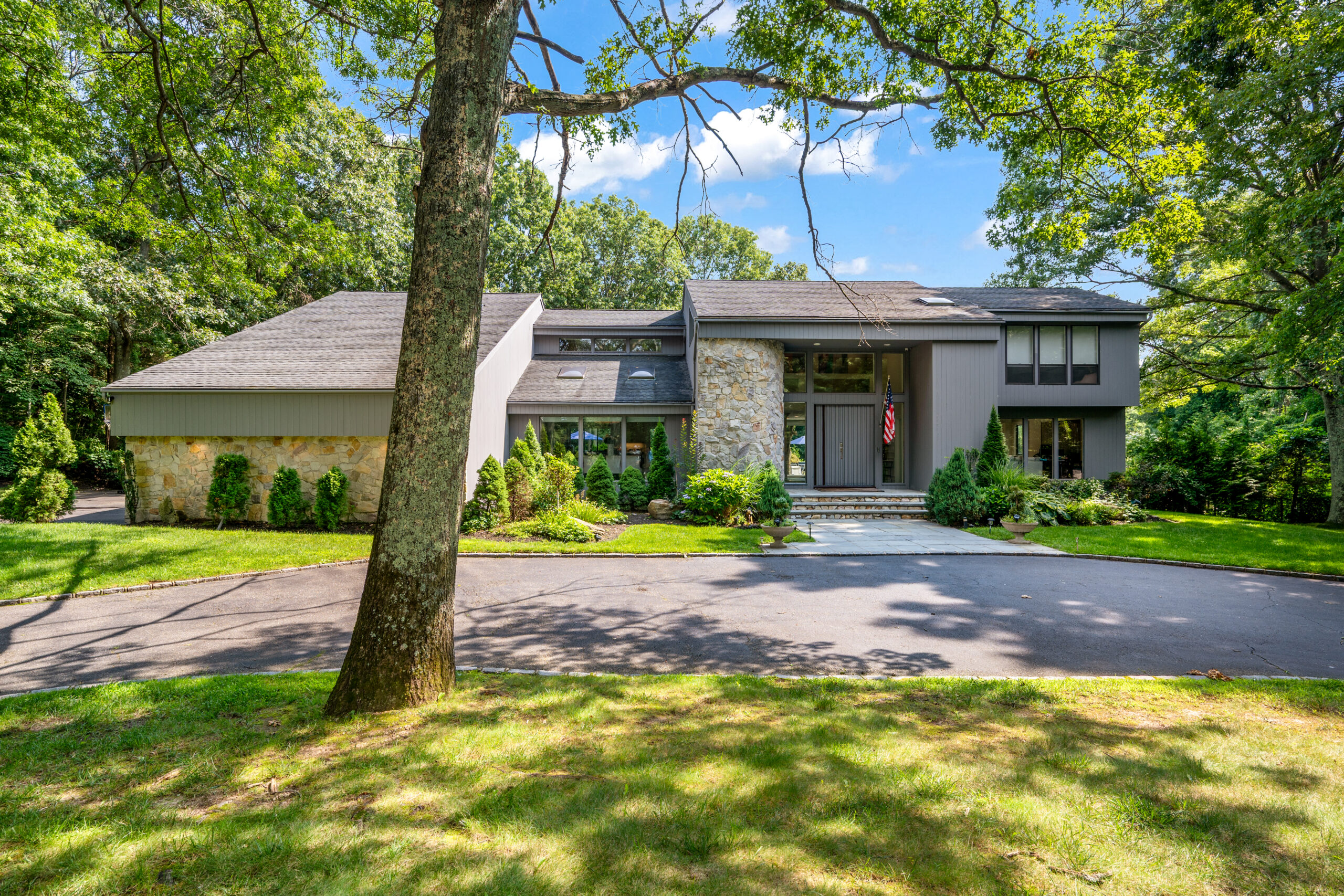 A split-level house with stone and gray siding, featuring a front door with an American flag, set behind a circular driveway surrounded by mature trees during the November 2026 a-list season.