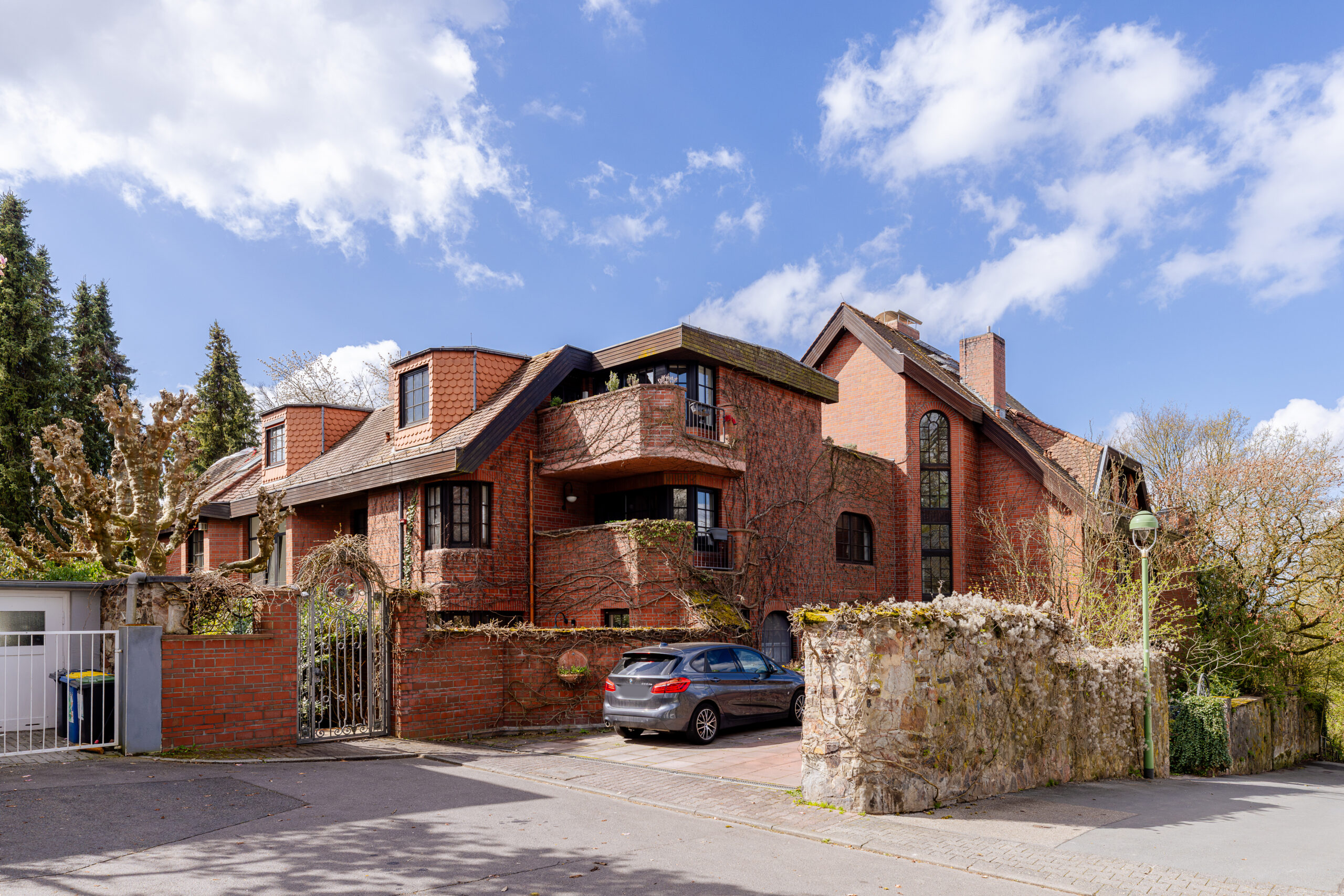 A multi-story red brick apartment building with gabled roofs and balconies, viewed in November 2026, with a grey car parked in the driveway.