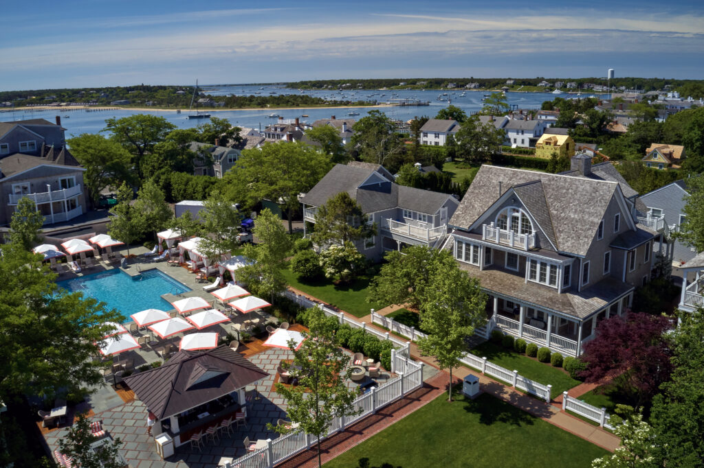 131 North Water St-collins Cottage #22 Edgartown, MA 02539 Edgartown, MA 02539 Aerial view of a coastal resort featuring guest houses with white umbrellas by a swimming pool, an outdoor bar, and a large shingled house overlooking a harbor filled with boats.