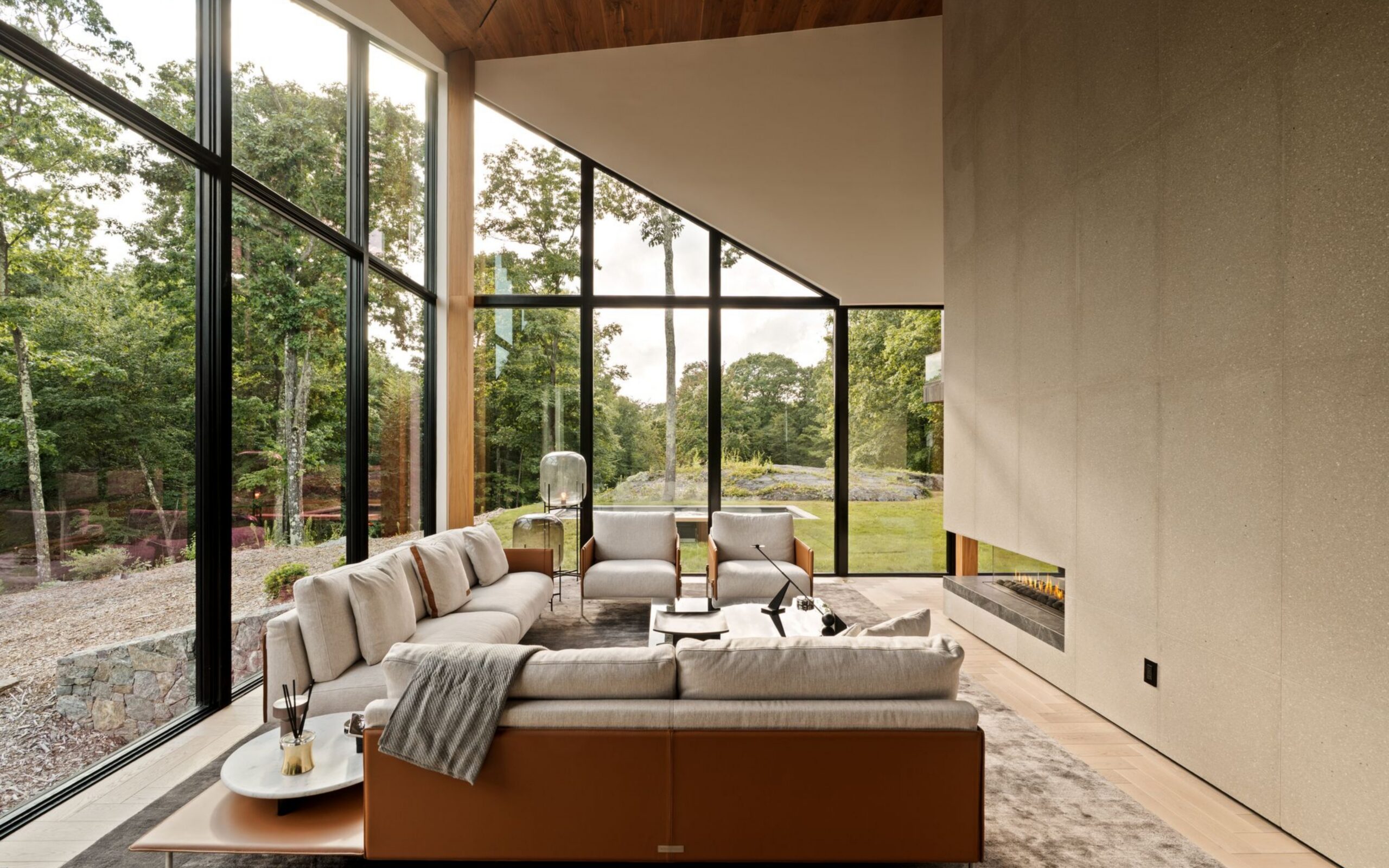 Modern living room in November 2026 with floor-to-ceiling windows, a large light-colored sectional sofa, two armchairs, and a built-in fireplace against a stone feature wall.