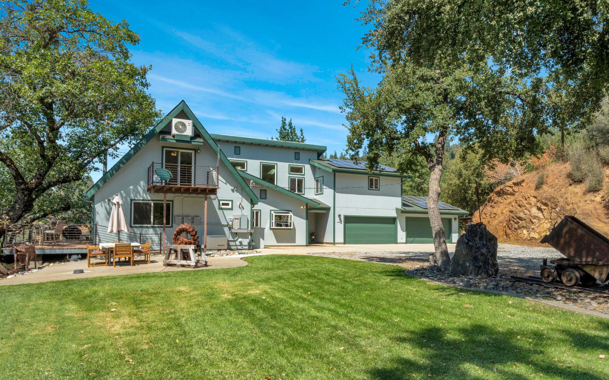 A two-story light blue house with a green roof in the Sierra Gold Country, featuring a two-car garage and a patio with a dining set in a grassy yard.