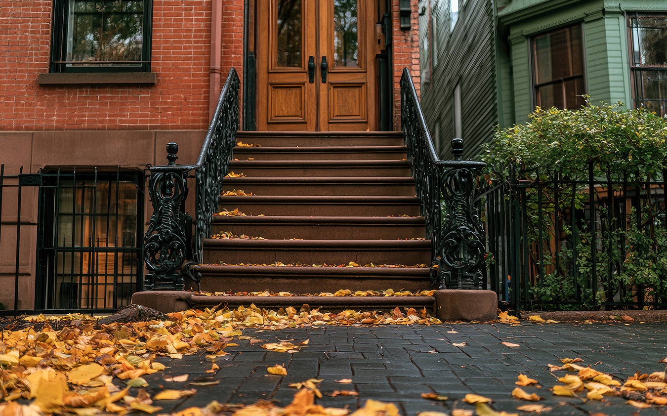A stoop with brown stairs and black iron railings leading to a wooden door, surrounded by fallen autumn leaves.