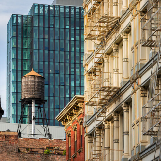 A historic building with external metal fire escapes stands next to a modern Manhattan condo skyscraper, with a rooftop water tower and a vintage street lamp in the foreground.