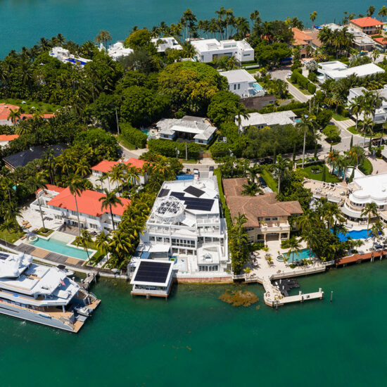 Aerial view of a South Florida real estate coastal neighborhood featuring large villas, swimming pools, and two yachts docked or cruising in the turquoise water.