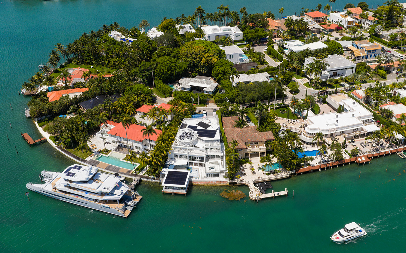 Aerial view of a South Florida real estate coastal neighborhood featuring large villas, swimming pools, and two yachts docked or cruising in the turquoise water.