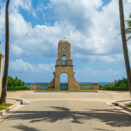 A stone clock tower stands at the end of a palm-lined path overlooking the ocean in a South Florida real estate setting.