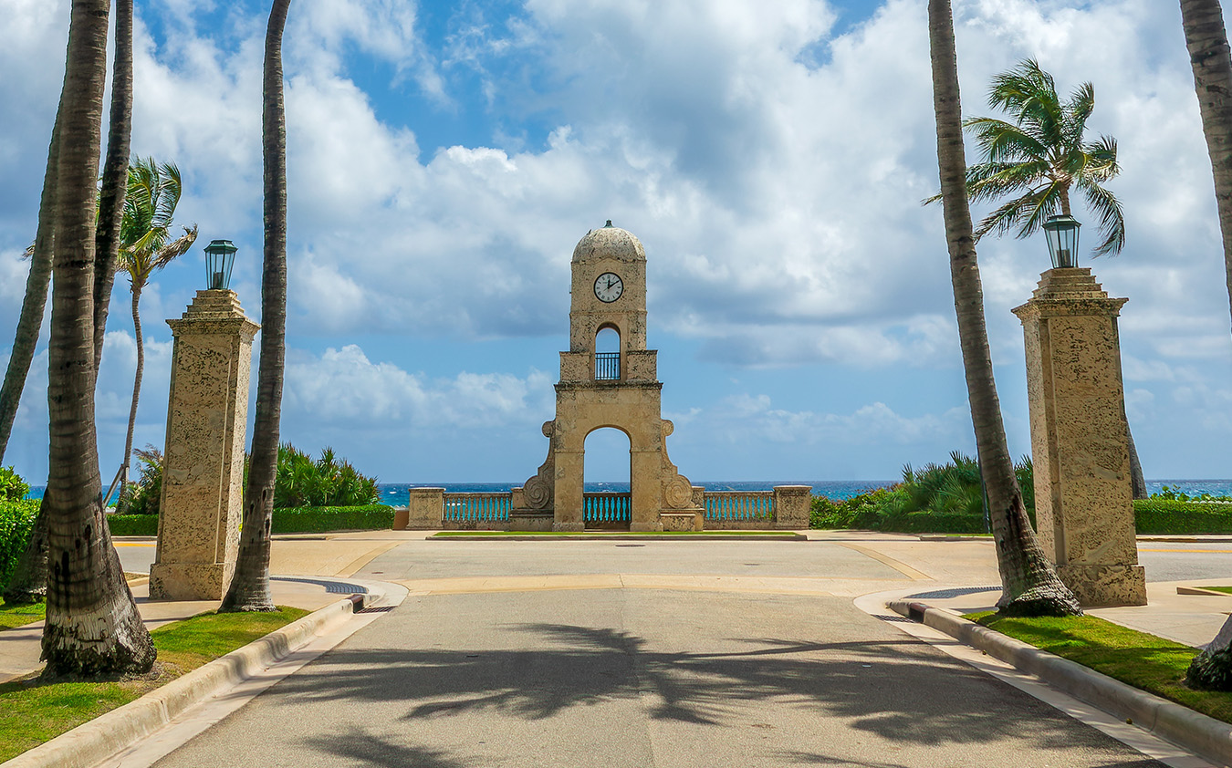 A stone clock tower stands at the end of a palm-lined path overlooking the ocean in a South Florida real estate setting.