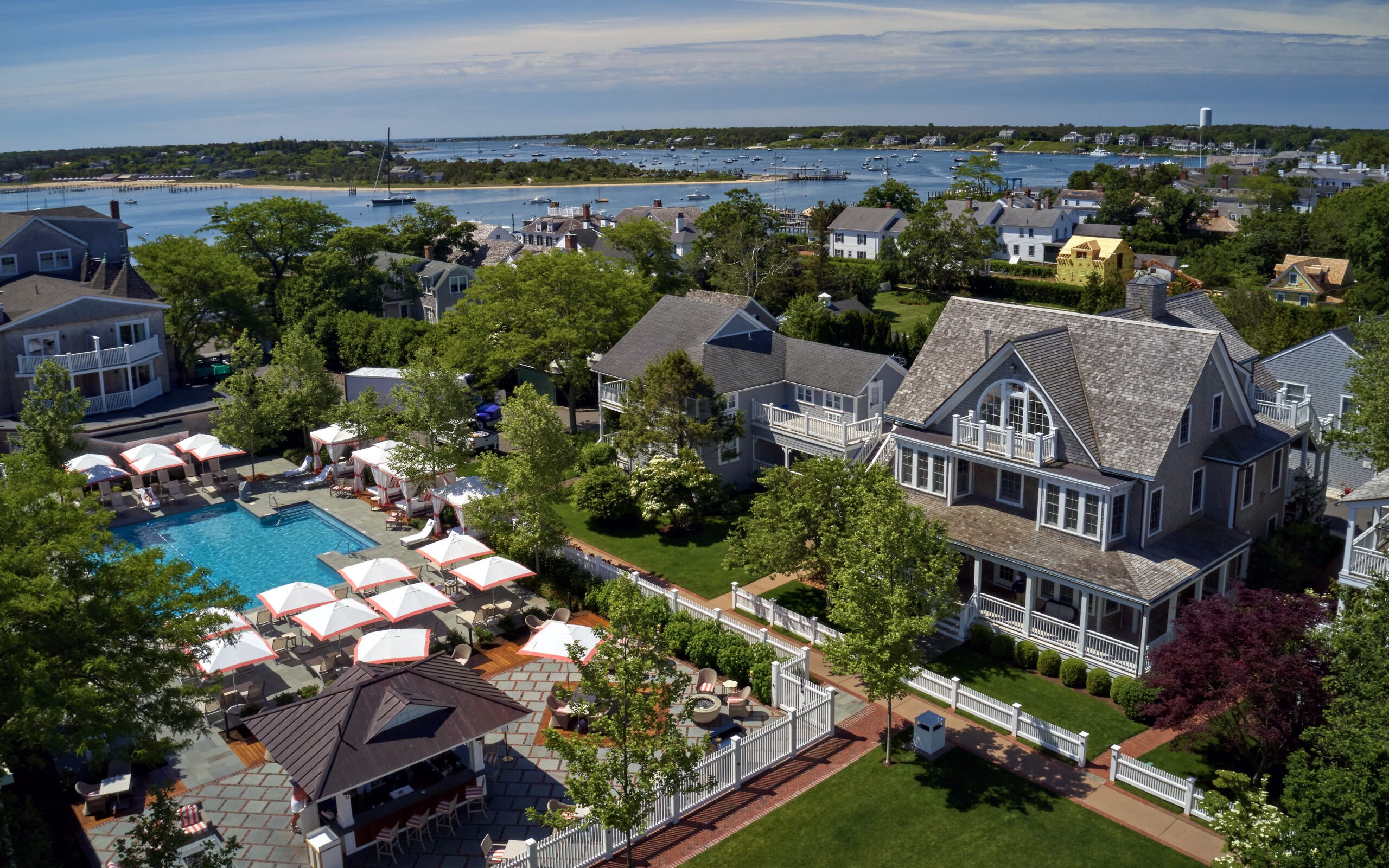 Aerial view of a coastal hotel-branded property featuring a swimming pool with lounge chairs and umbrellas, adjacent to shingled houses and a harbor with boats in the distance.