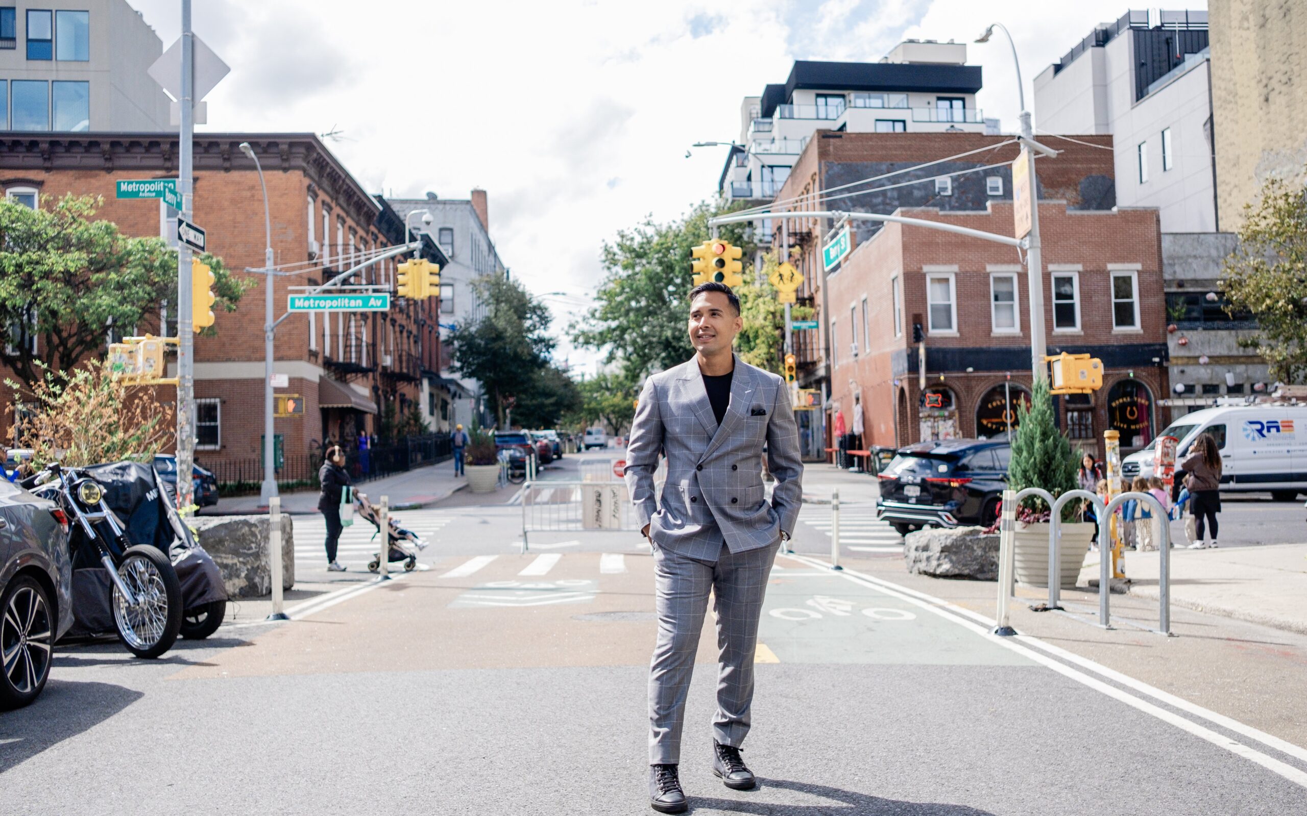 A man in a grey plaid suit stands in the middle of a city intersection with street signs reading Metropolitan Avenue.
