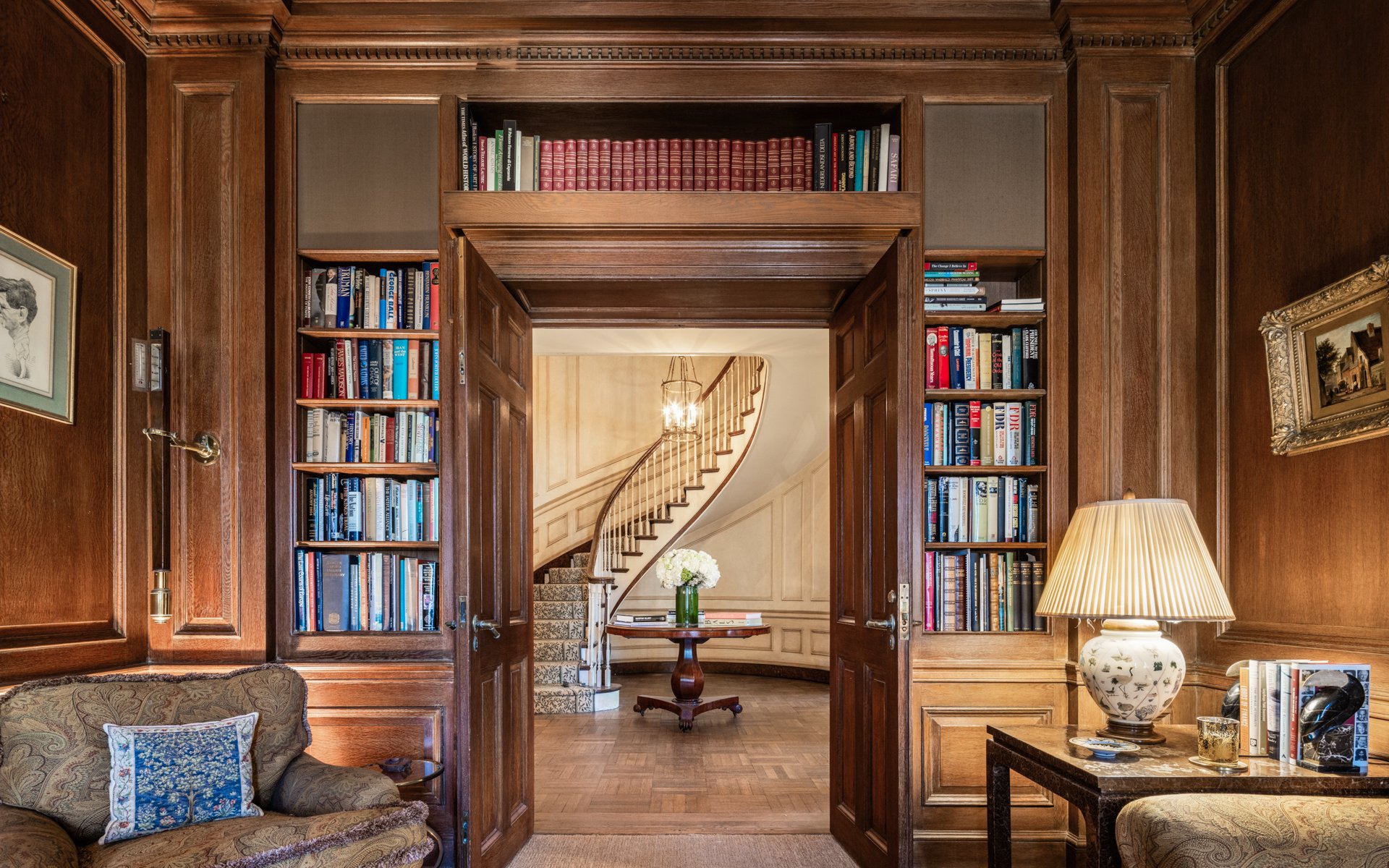 A view through a paneled wood doorway into a foyer with a curved staircase, flanked by built-in bookshelves in a home office library.