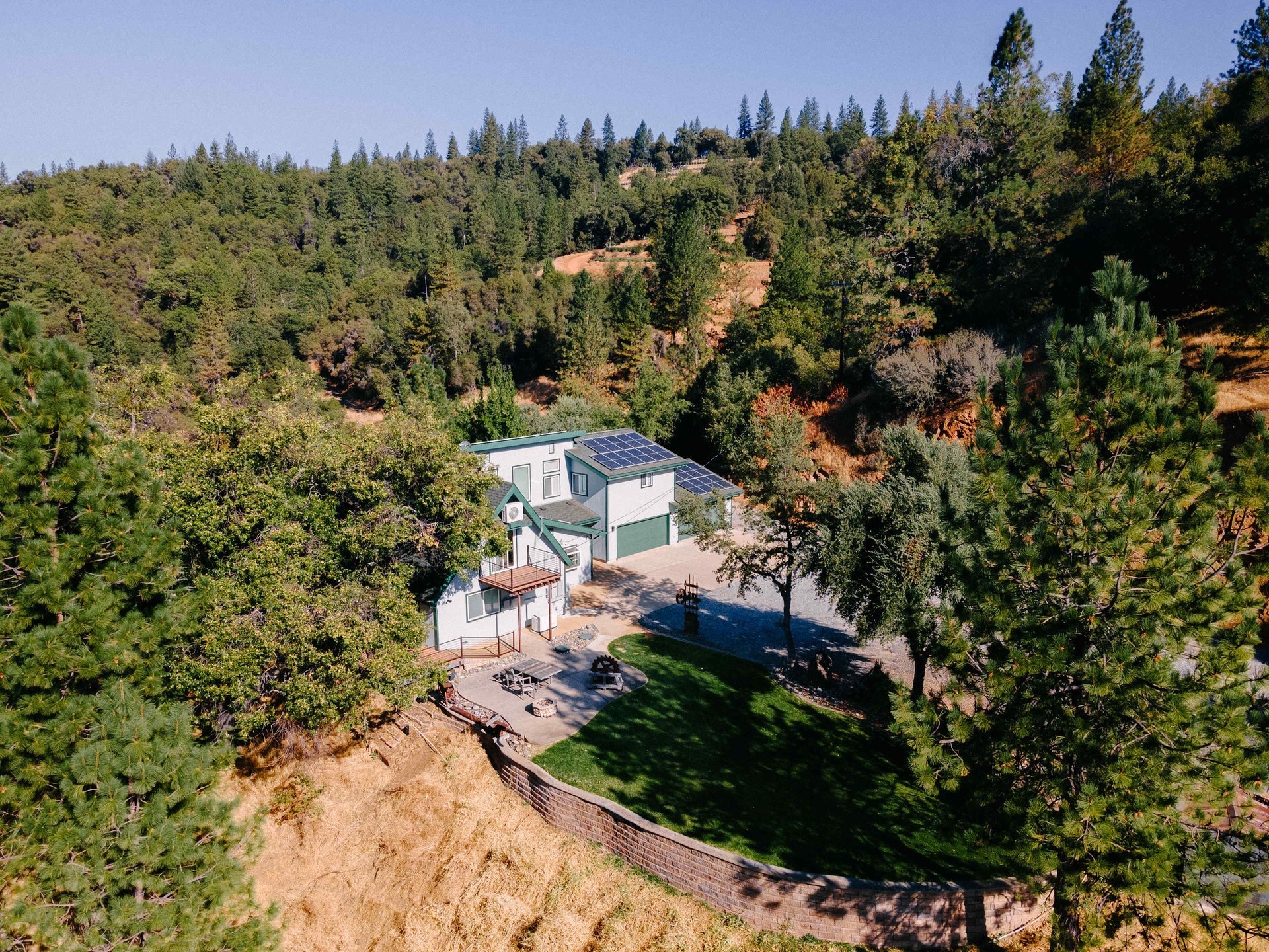 Aerial view of a house with solar panels on the roof, surrounded by a forest and a terraced lawn with a patio.